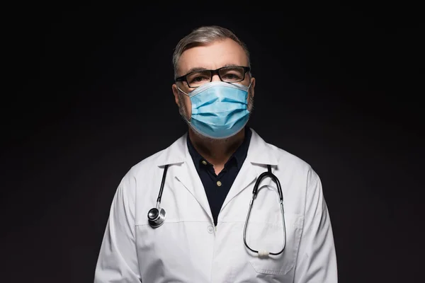 Senior doctor in white coat and medical mask looking at camera isolated on black — Stock Photo