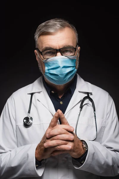 Doctor in medical mask and eyeglasses standing with clasped hands and looking at camera isolated on black — Foto stock