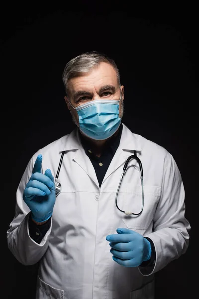 Senior doctor in medical mask and latex gloves pointing with finger while looking at camera isolated on black — Stock Photo