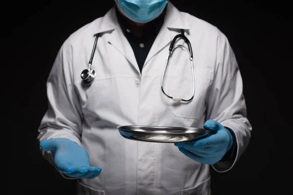 Cropped view of doctor in medical mask and latex gloves holding stainless medical tray isolated on black — Photo de stock