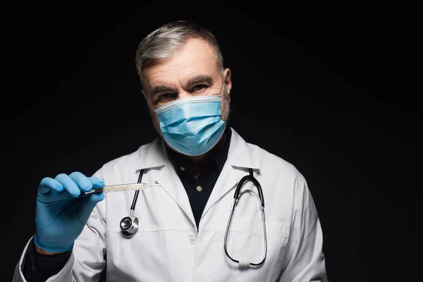 Doctor in medical mask and white coat holding thermometer isolated on black — Stock Photo