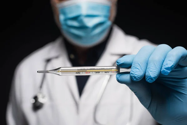 Partial view of blurred doctor in medical mask holding thermometer isolated on black — Stockfoto