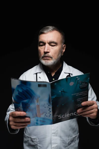 Bearded physician in white coat reading science magazine isolated on black — Stock Photo
