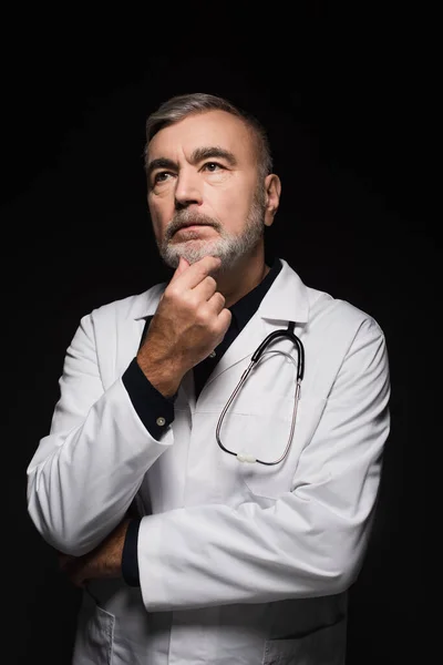 Pensive doctor touching chin and looking away while thinking isolated on black — Stock Photo
