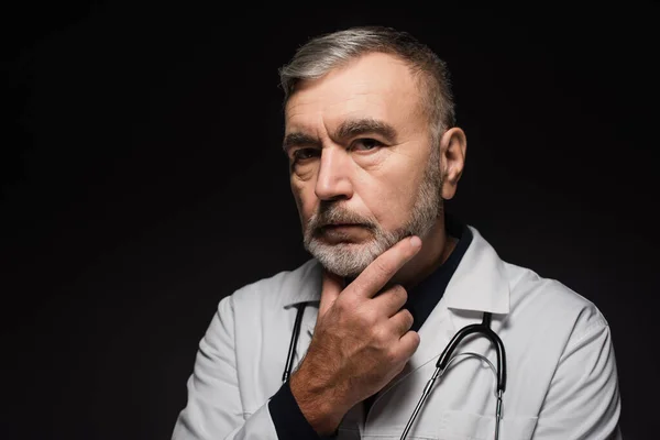 Pensive bearded doctor looking at camera while touching chest isolated on black — Stock Photo
