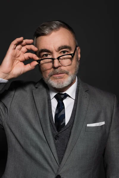 Businessman in stylish formal wear adjusting eyeglasses and looking at camera isolated on dark grey — Fotografia de Stock