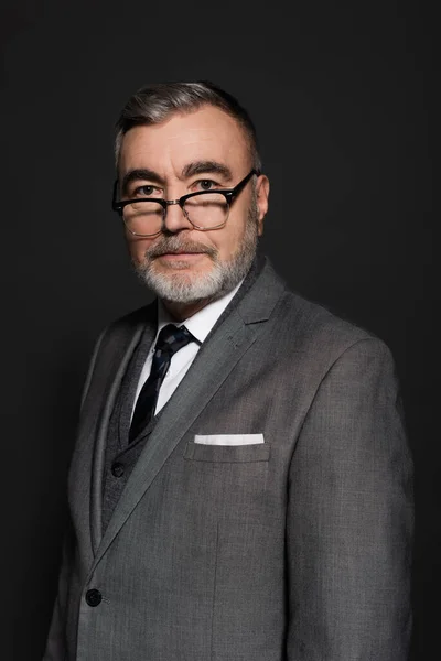 Senior businessman in formal wear and eyeglasses looking at camera isolated on dark grey — Photo de stock