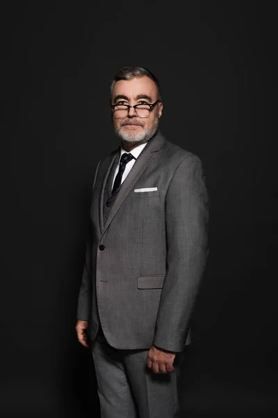 Bearded senior man in suit and eyeglasses looking at camera on dark grey — Foto stock