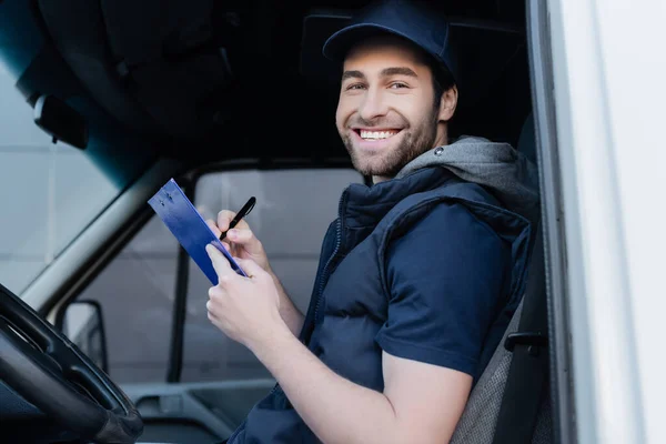 Cheerful delivery man holding clipboard and pen in car — стоковое фото