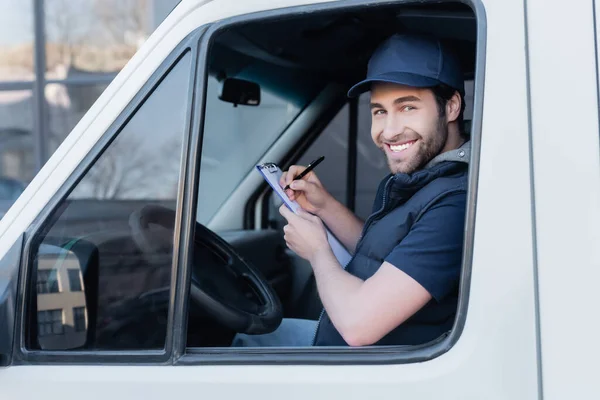 Young courier smiling at camera while writing on clipboard in car — Photo de stock