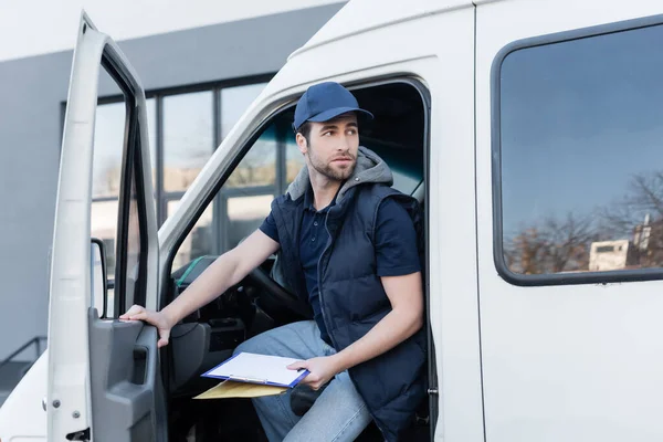 Young delivery man holding parcel and clipboard while sitting in car — стоковое фото