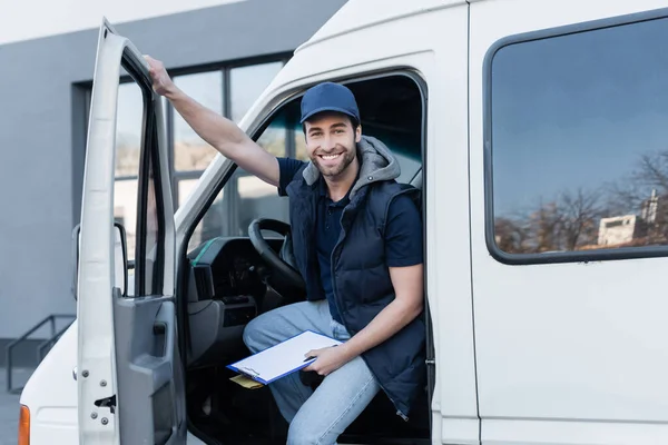 Happy delivery man looking at camera while holding clipboard and parcel in auto — Photo de stock