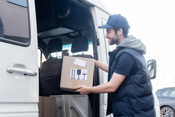 Side view of smiling courier in uniform taking package from car outdoors - foto de stock