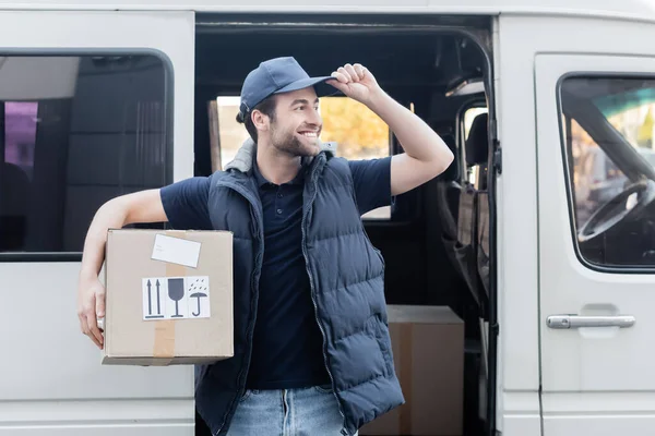 Positive courier holding carton box and looking away near auto outdoors — стоковое фото