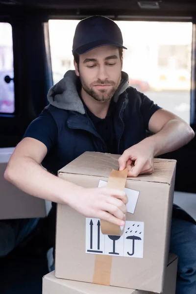 Чоловік-доставка, який наносить клейку стрічку на картонну коробку в авто — стокове фото
