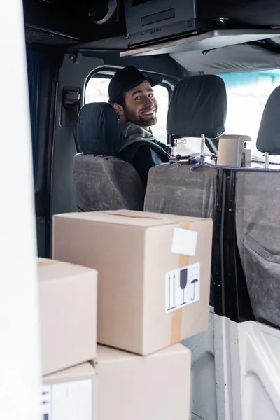 Smiling delivery man looking at camera near packages in car — Stock Photo