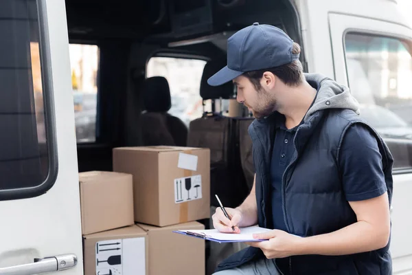 Side view of courier writing on clipboard near blurred boxes in car — Fotografia de Stock