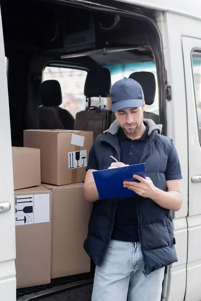 Young courier in uniform writing on clipboard near boxes with signs in auto — Stock Photo