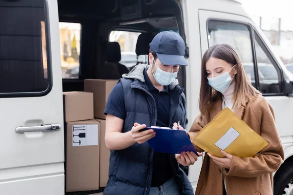 Delivery man in medical mask holding clipboard near woman with parcel and car outdoors — Stock Photo