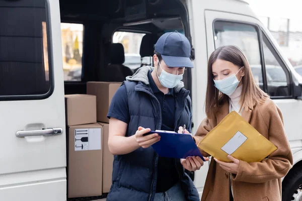 Delivery man in medical mask holding clipboard near woman with parcel and car outdoors — стоковое фото