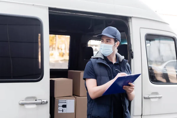 Courier in medical mask writing on clipboard near boxes in car outdoors — стоковое фото