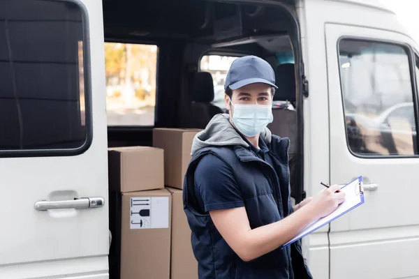 Delivery man in medical mask writing on clipboard near carton boxes in car outdoors — Stock Photo