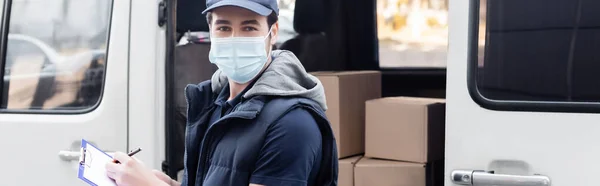 Courier in protective mask holding clipboard and looking at camera near auto on urban street, banner — стокове фото