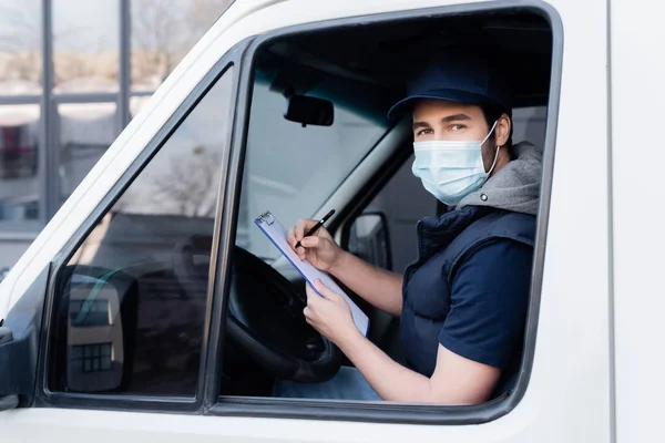 Courier in medical mask holding clipboard and looking at camera in car — стокове фото