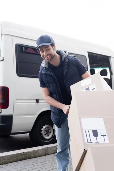 Smiling courier looking at carton boxes with signs near auto outdoors — Stock Photo