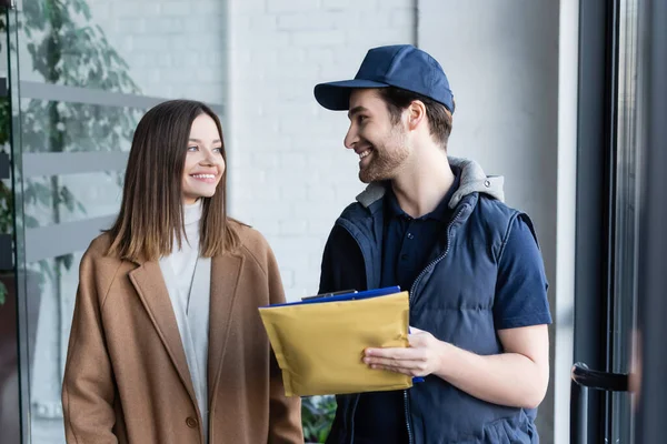 Smiling courier holding clipboard and parcel near woman in hallway - foto de stock