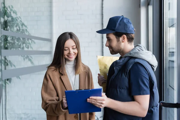 Smiling woman looking at clipboard near courier with parcel in hallway — стоковое фото