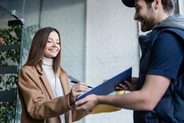 Smiling woman with parcel writing on clipboard near blurred courier in hallway — Stockfoto