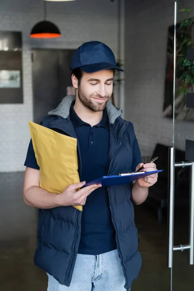 Positive delivery man holding parcel and writing on clipboard in hallway — стоковое фото