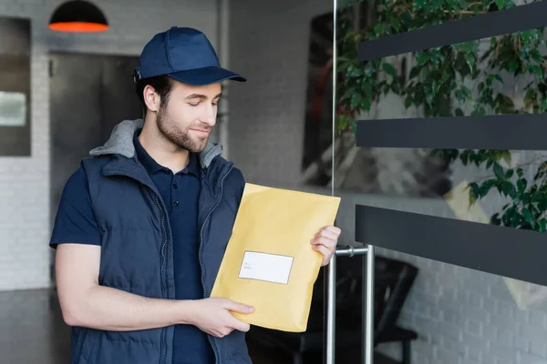 Молодий чоловік доставки в уніформі тримає посилку з наклейкою в коридорі — стокове фото