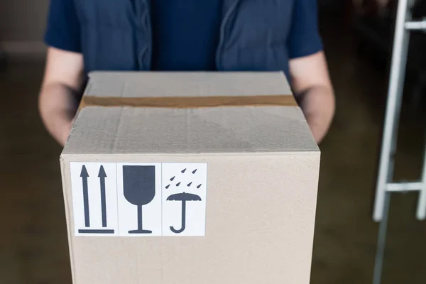 Cropped view of courier holding carton box with signs in hallway — Stock Photo