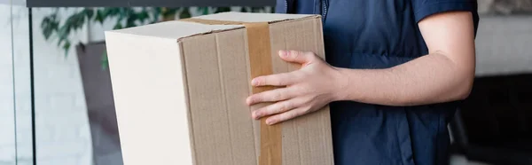 Cropped view of delivery man holding carton box in hallway, banner — Photo de stock