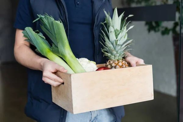 Cropped view of courier holding wooden box with fresh food in hallway - foto de stock