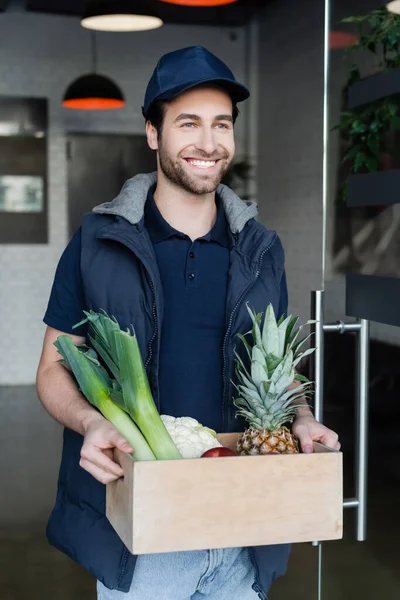 Smiling delivery man holding wooden box with food in hallway — Stock Photo