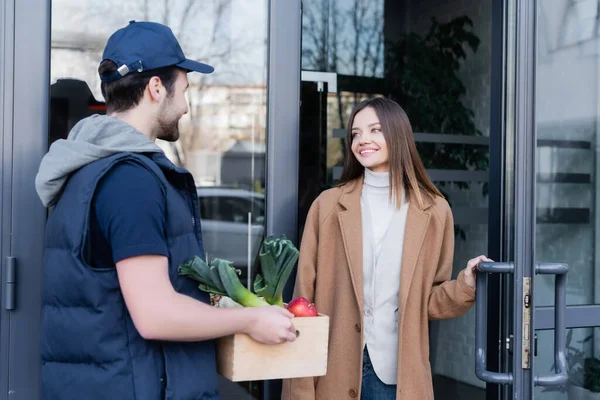 Smiling woman looking at courier with fresh food near door of building outdoors — Stock Photo