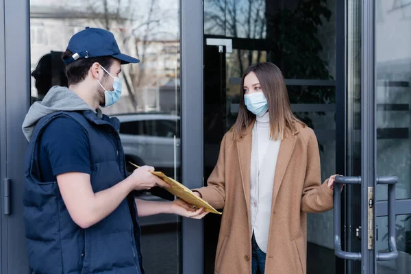 Жінка в медичній масці бере посилку від кур'єра біля дверей будівлі на відкритому повітрі — стокове фото