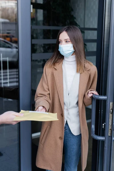 Delivery man holding parcel near woman in medical mask and door of building outdoors — Foto stock