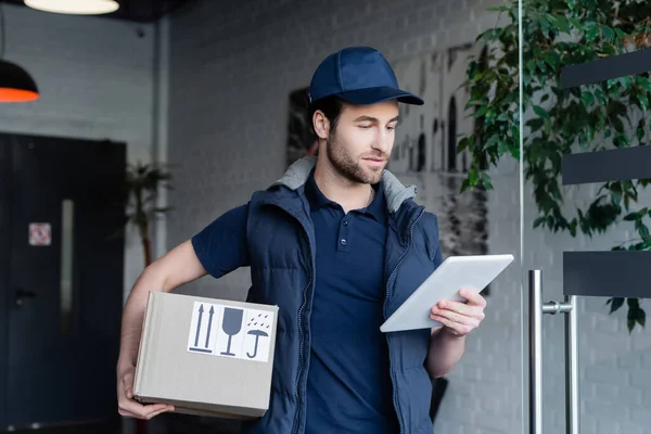 Delivery man in uniform holding carton box and using digital tablet in hallway — стоковое фото