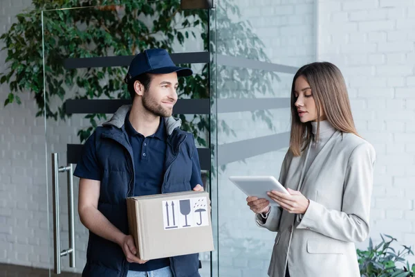 Young woman holding digital tablet near smiling delivery man with carton box in hallway — Stockfoto