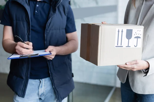 Cropped view of courier writing on clipboard near girlfriend with cardboard box in hallway — Stock Photo