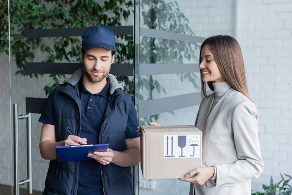 Smiling woman holding carton box with symbols near delivery man writing on clipboard in hallway — Foto stock