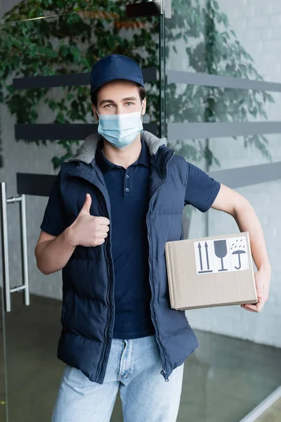 Courier in medical mask holding box with symbols and showing like in hallway — стоковое фото
