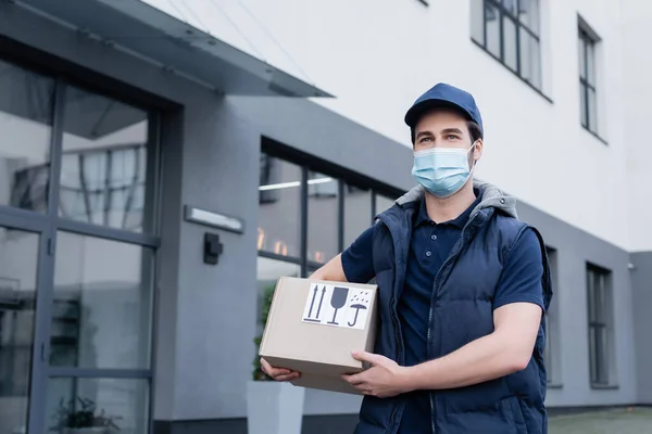 Delivery man in medical mask holding carton box with signs on urban street — Foto stock