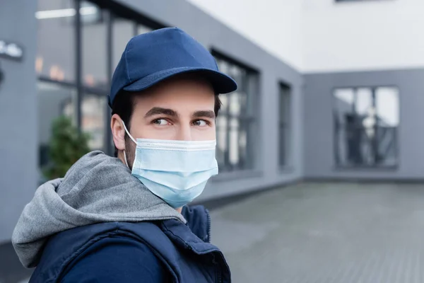 Young delivery man in medical mask and cap looking away on urban street — стоковое фото