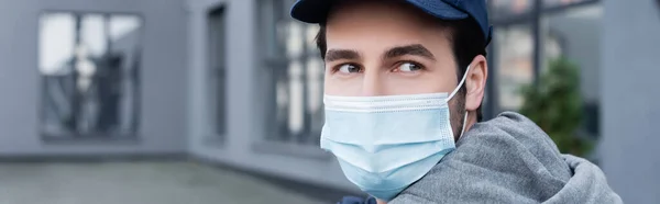 Courier in medical mask and cap looking away on urban street, banner — Stockfoto