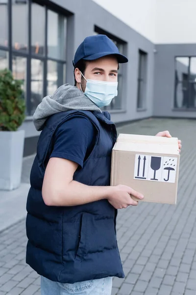 Young delivery man in medical mask looking at camera and holding box with signs on urban street — Stockfoto